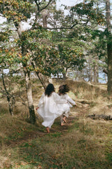 Two women in white dresses walking through a forest path.
