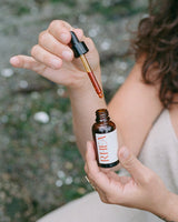 Person holding a dropper with a bottle labeled RHEA against a blurred natural green background